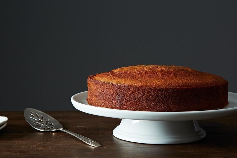 An olive oil cake sits on a white cake stand on a wooden surface. It is an unadorned, golden-brown cake.